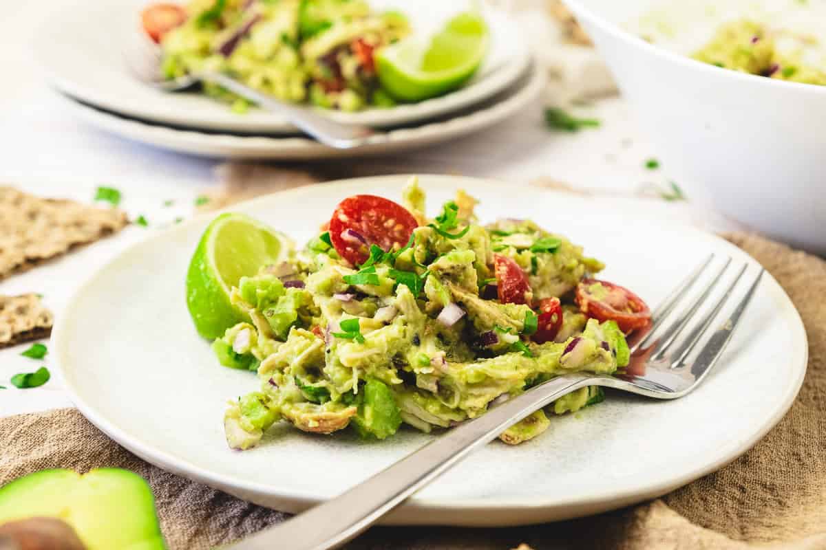 A plate of guacamole made with fresh avocado, cherry tomatoes, red onion, and lime, served with a fork on a white plate—perfect as a vibrant side for your favorite chicken salad recipe.