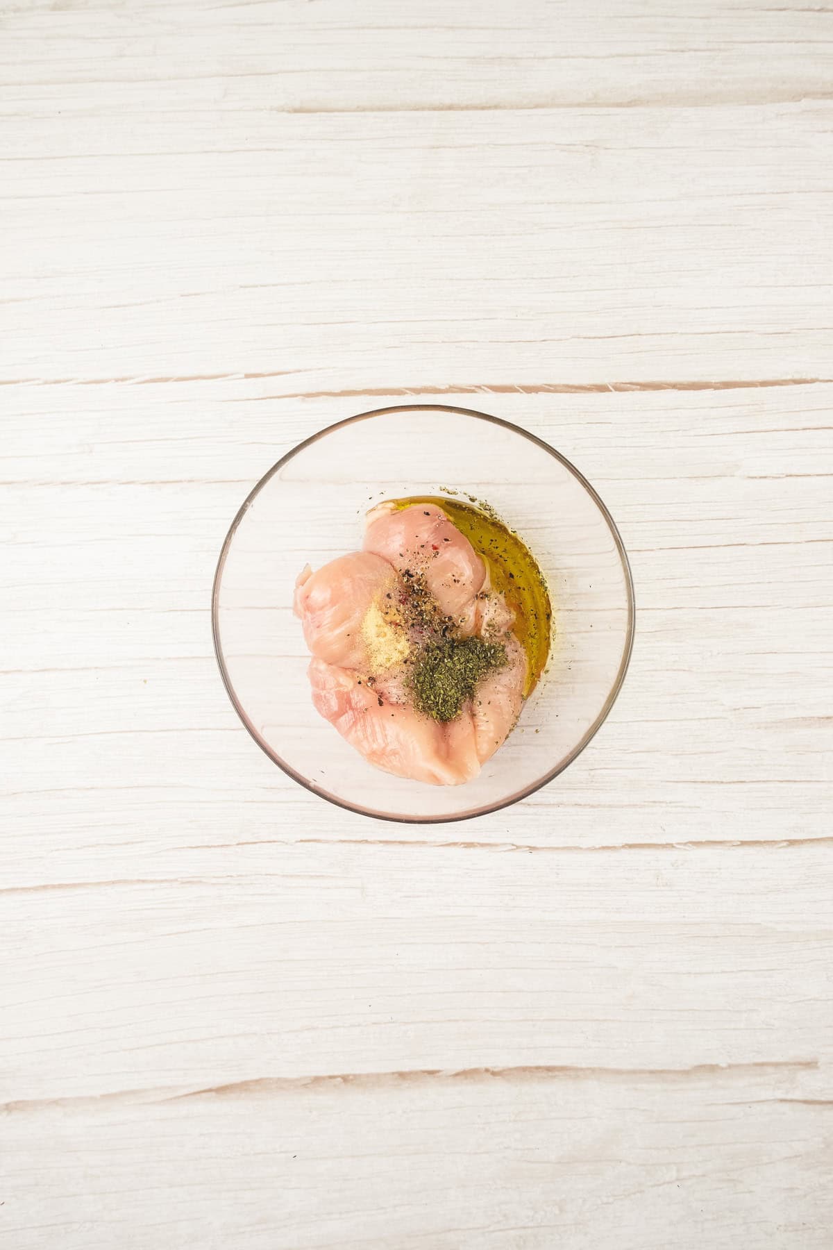 Raw chicken breast with spices and oil in a glass bowl on a white wooden surface, perfect for your next chicken salad recipe.