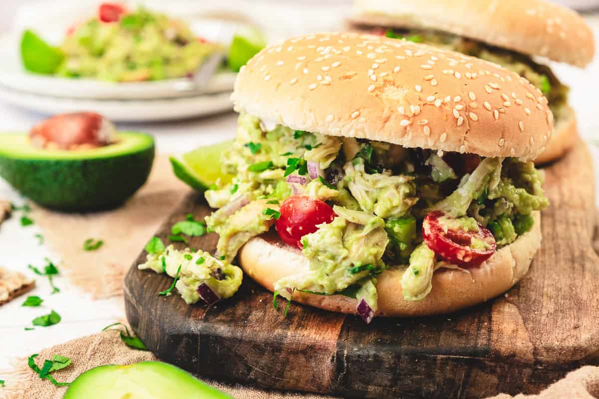 A sandwich bun filled with guacamole, shredded chicken salad, tomatoes, and herbs on a wooden board—perfect for trying a new recipe.