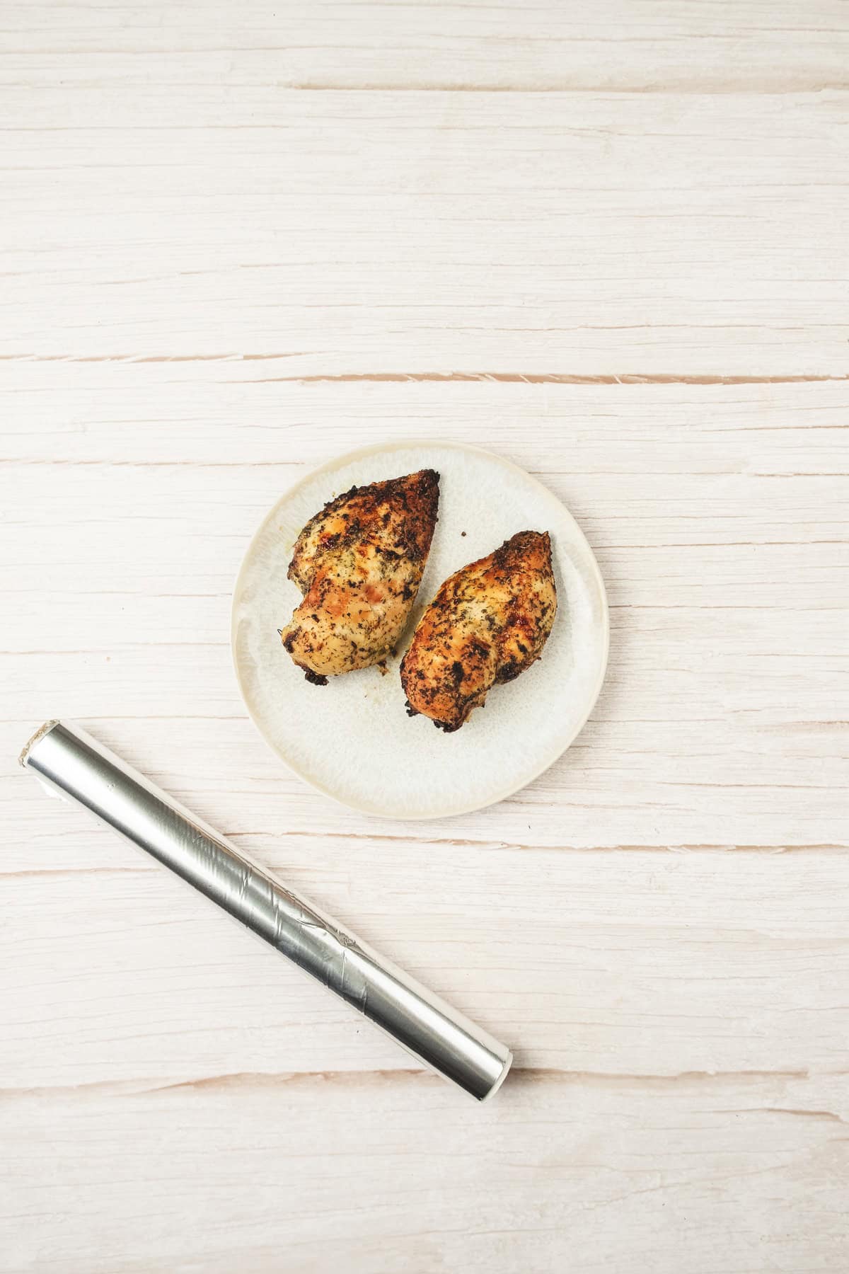 A small plate with two pieces of grilled chicken beside a silver cylindrical object on a light wooden surface—perfect for starting your next chicken salad recipe.