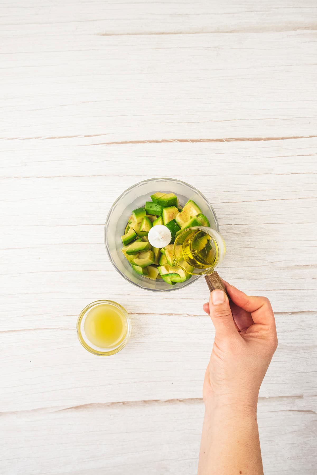 A hand pours olive oil into a food processor with chopped avocado and jalapeño on a white surface, prepping the perfect base for a creamy avocado chicken salad recipe.