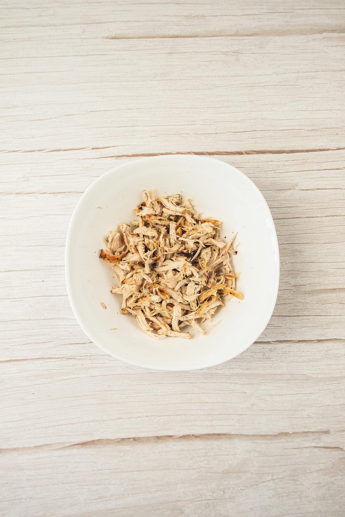 Shredded cooked chicken in a white bowl on a light wooden surface, perfect for your next chicken salad recipe.