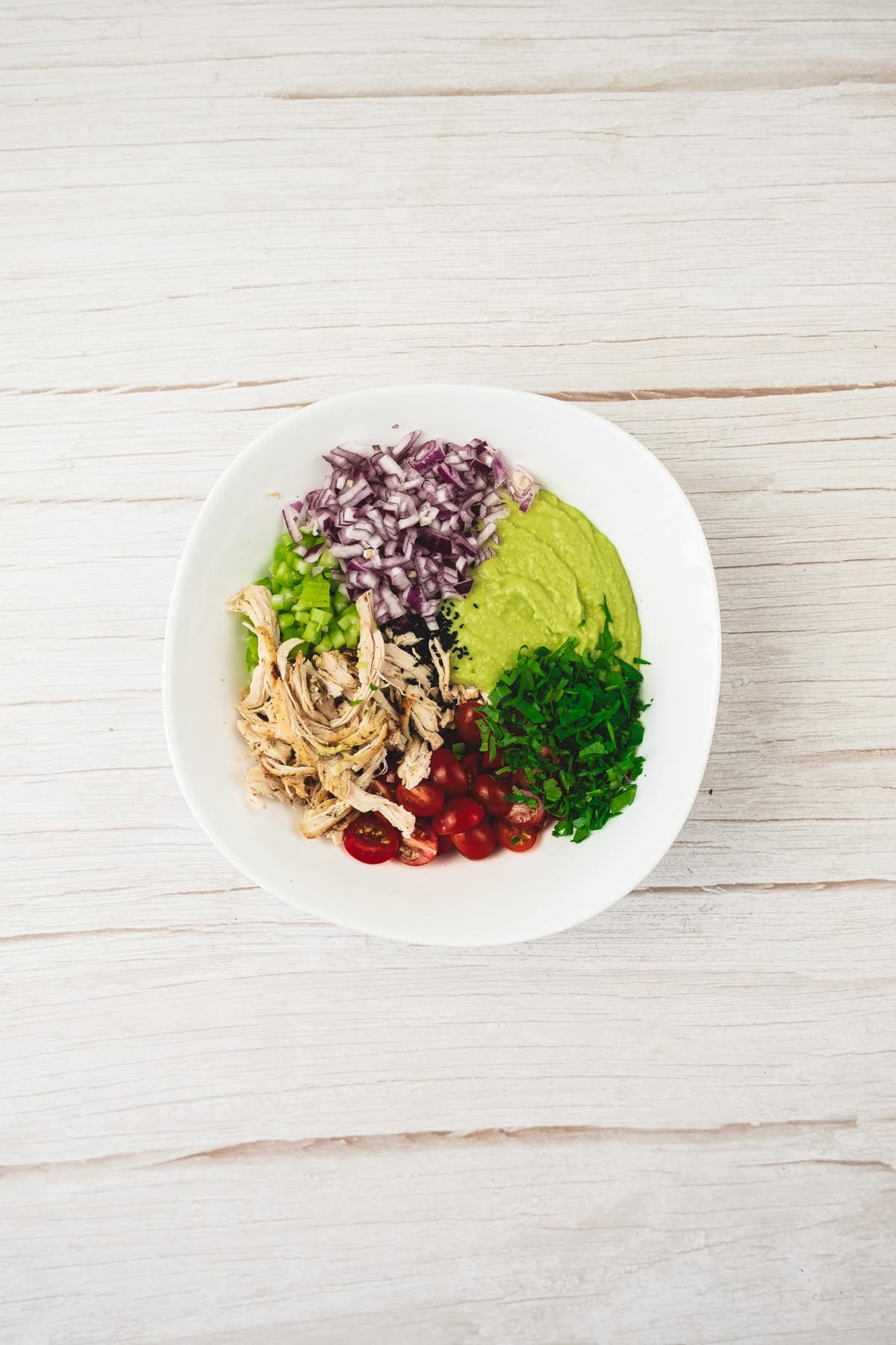 Bowl with shredded chicken, diced red onion, cherry tomatoes, green herbs, and creamy avocado guacamole—perfect for a fresh chicken salad recipe on a white surface.