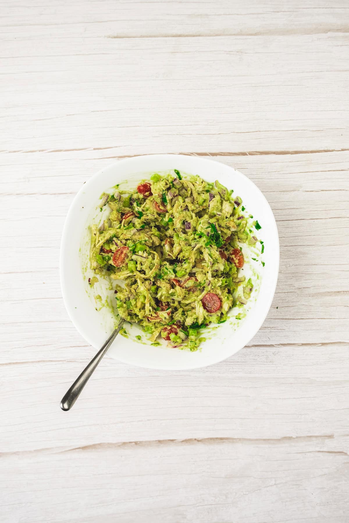 A bowl of guacamole made with fresh avocado, tomato, and onion sits with a spoon on a light wooden surface.