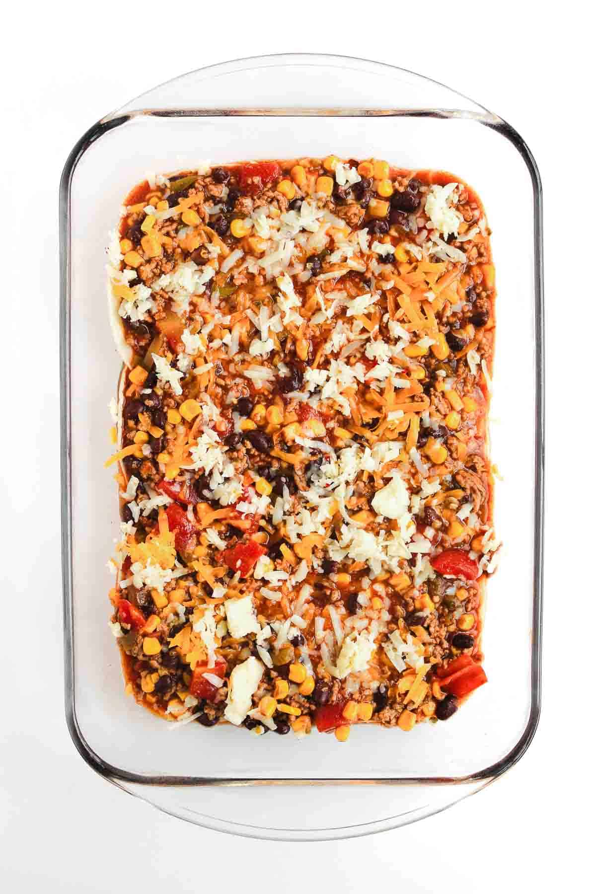 A glass baking dish with a layered Beef Enchilada Casserole, topped with cheese, beans, corn, and vegetables&mdash;a delicious casserole recipe perfect for any gathering.