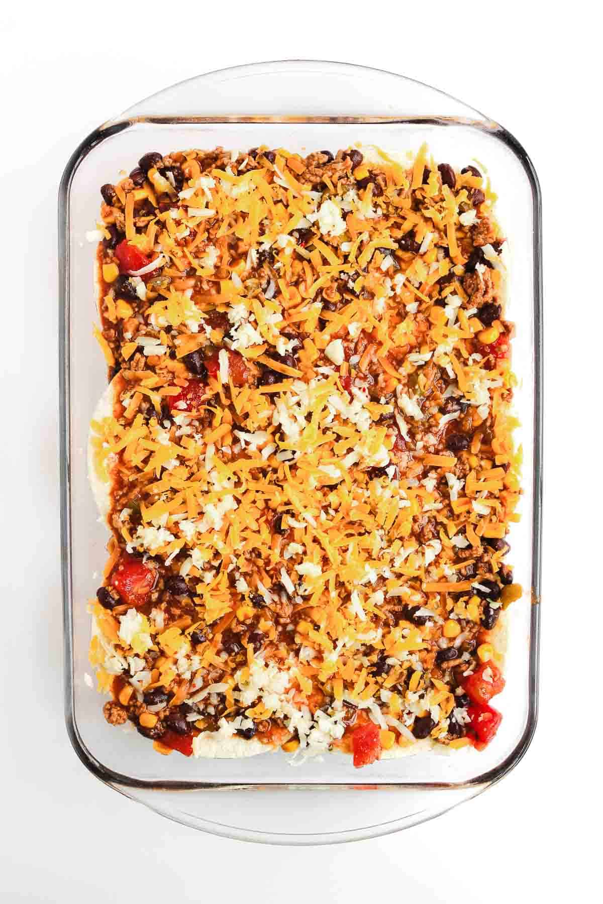 A glass baking dish filled with a layered Beef Enchilada Casserole, topped with shredded cheese, beans, and tomatoes&mdash;a flavorful casserole recipe perfect for dinner.