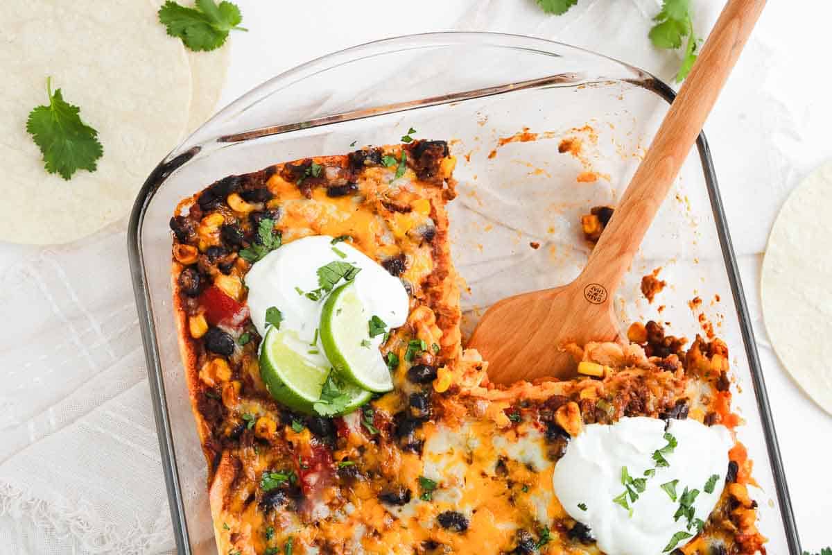 A glass baking dish with a cheesy Beef Enchilada Casserole, topped with sour cream, lime slices, and chopped cilantro.