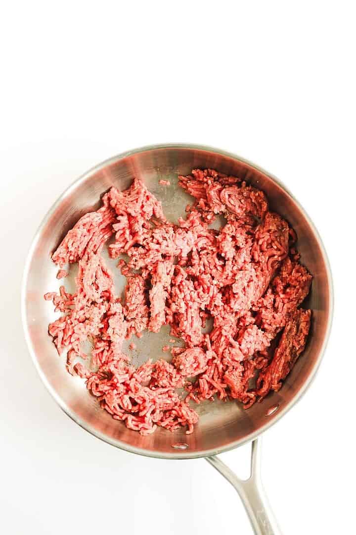 Raw ground beef in a stainless steel skillet on a white background, perfect for starting your favorite casserole recipe like a classic Beef Enchilada Casserole.