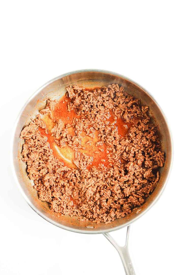 Ground beef with savory sauce simmers in a stainless steel pan&mdash;perfect for a classic Beef Enchilada Casserole recipe&mdash;set on a white background.