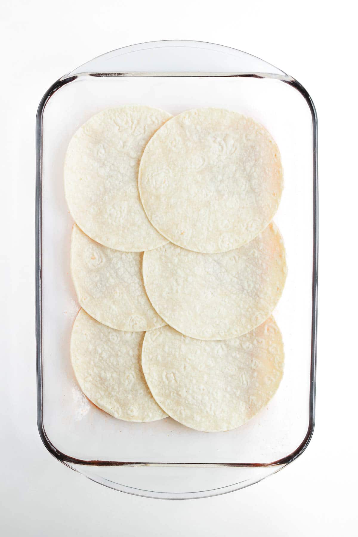 Six corn tortillas arranged flat in a glass baking dish, ready to become the base for a delicious Beef Enchilada Casserole recipe.