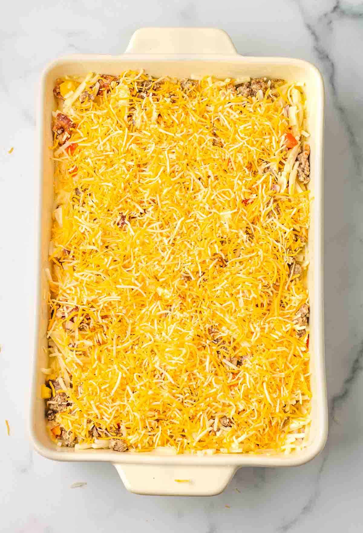 A Hashbrown Breakfast Casserole filled with layers and topped with shredded cheddar cheese sits on a marble surface, ready to serve.