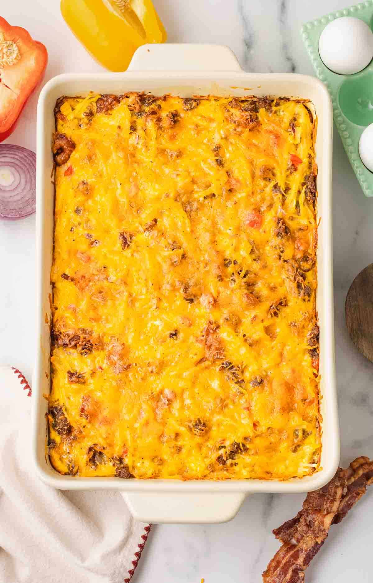 Baked hashbrown casserole with melted cheese in a rectangular dish, surrounded by eggs, bacon, and vegetables.