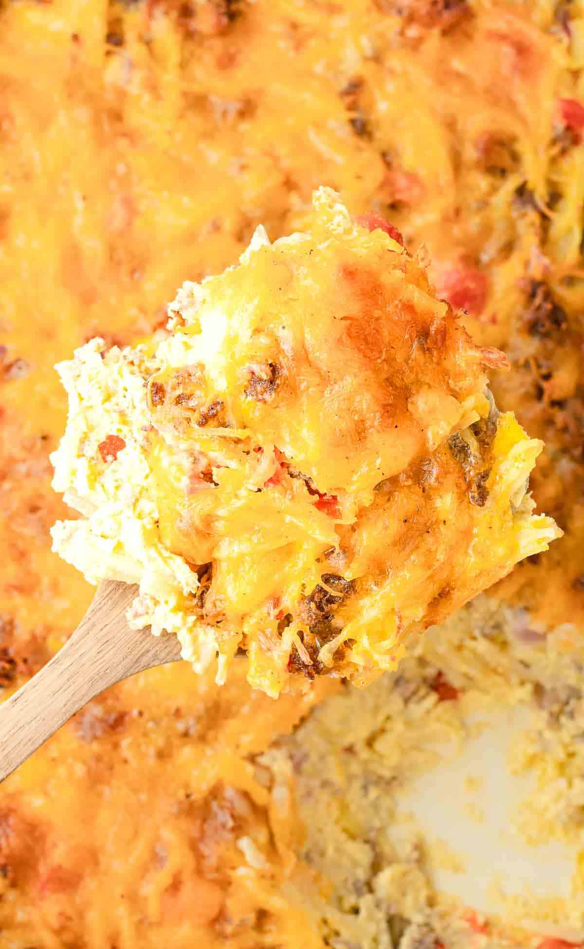 A wooden spoon holds a cheesy Hashbrown Breakfast Casserole with melted shredded cheese and bits of vegetables on top.