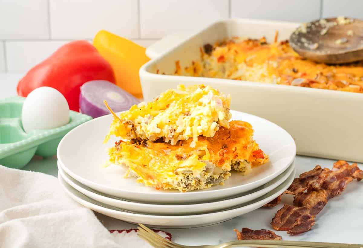 A slice of hashbrown breakfast casserole with eggs, cheese, and sausage sits on a stack of white plates; the casserole dish is in the background.