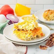 A slice of Hashbrown Breakfast Casserole on a plate with bacon, cheese, onion, and peppers in the background.