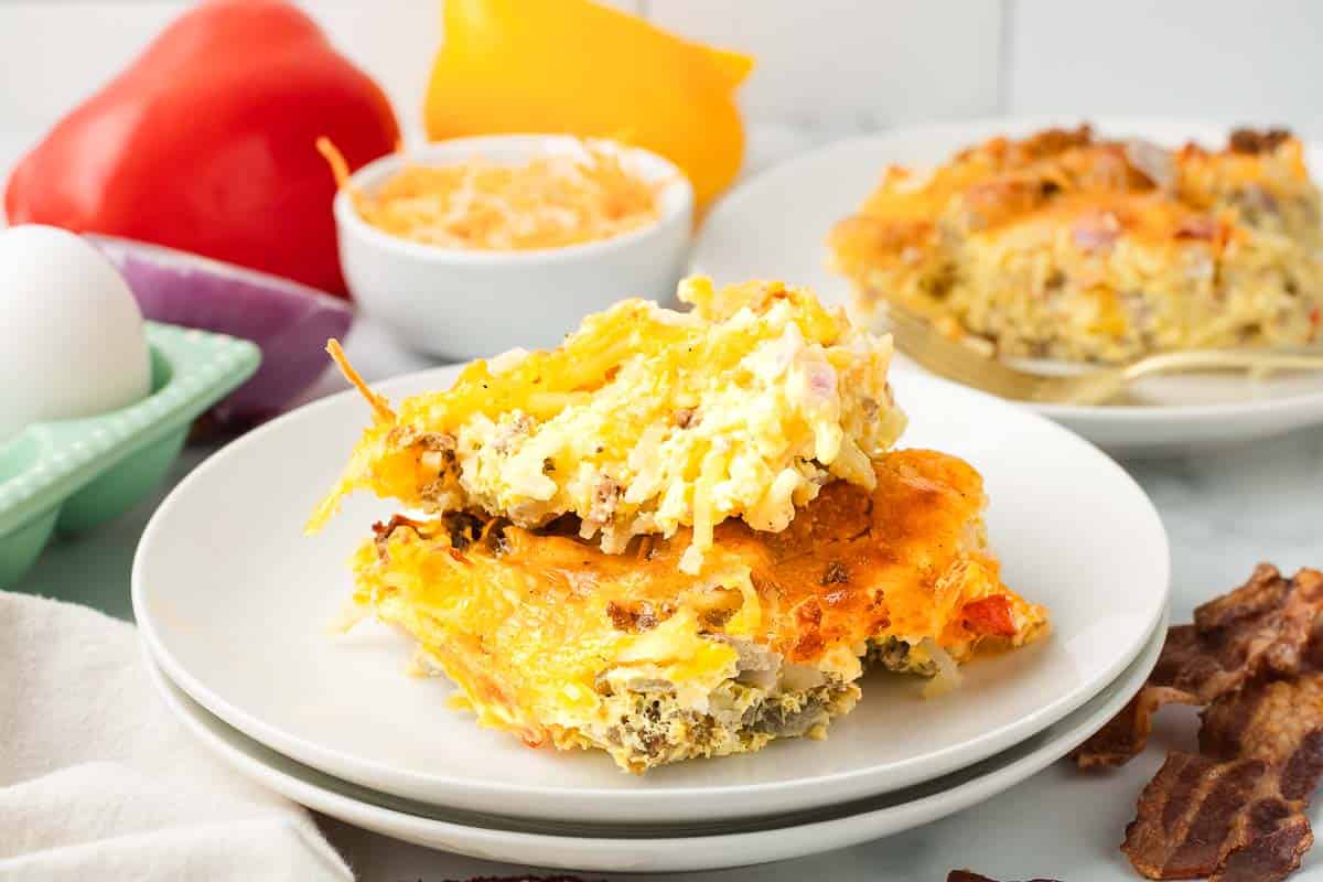 Two slices of cheesy hashbrown breakfast casserole on a white plate, with peppers, eggs, and bacon in the background.