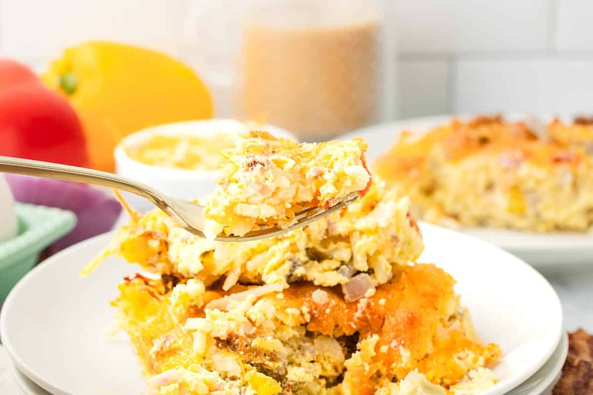 A fork holds a bite of cheesy Hashbrown Breakfast Casserole above a serving on a white plate.