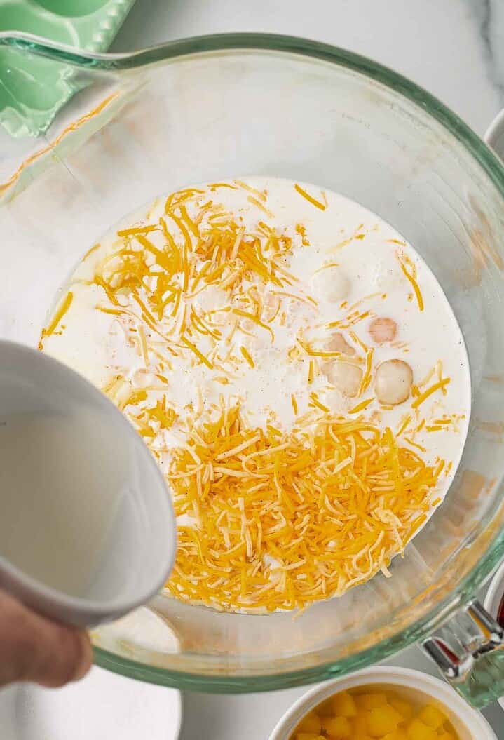 A bowl with shredded cheese, cream, and someone pouring liquid into the mixture from above&mdash;perfect for preparing a delicious Hashbrown Breakfast Casserole.