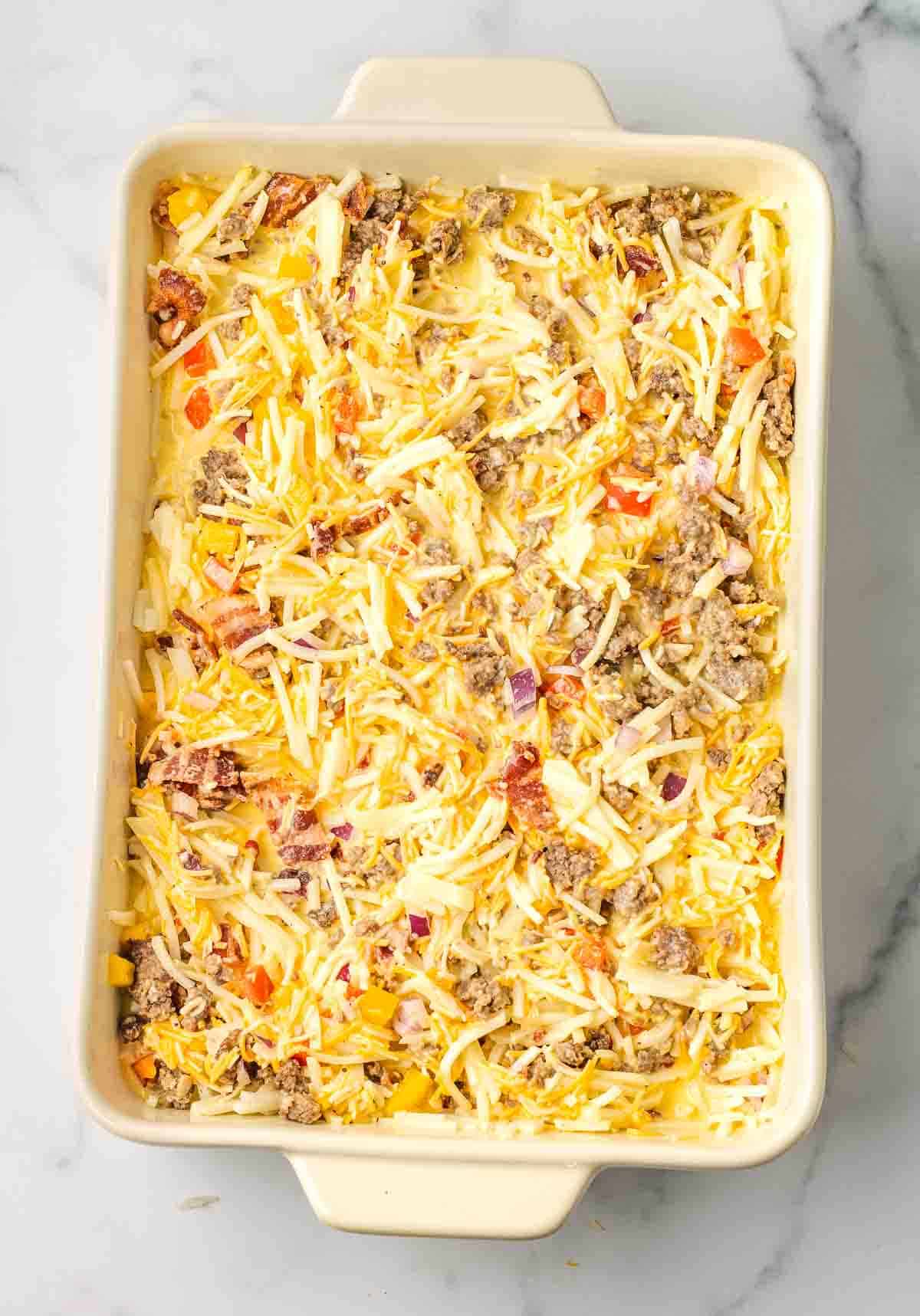 A Hashbrown Breakfast Casserole dish filled with an unbaked mixture of shredded cheese, sausage, bacon, and chopped vegetables.