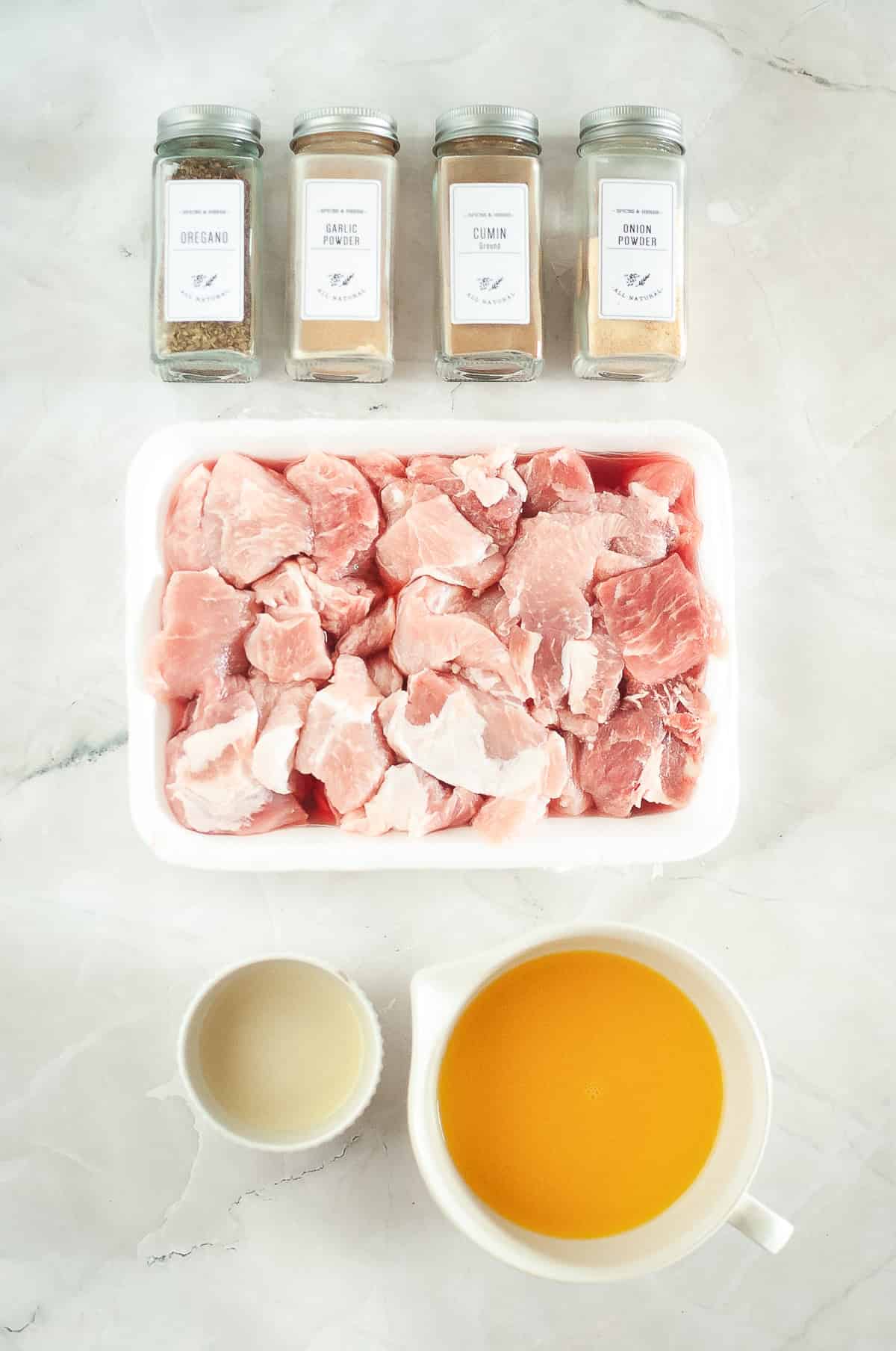 Raw pork chunks in a tray, four spice jars, a cup of vinegar, and a jug of orange juice on a marble surface&mdash;perfect for making flavorful Carnitas in your Instant Pot.