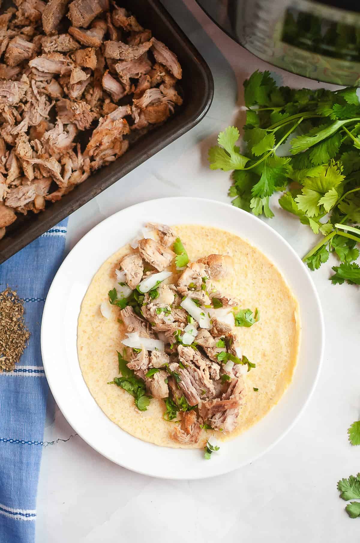 A tortilla topped with savory Instant Pot Carnitas, chopped onions, and cilantro on a white plate beside fresh cilantro&mdash;a delicious twist on the classic Carnitas recipe.