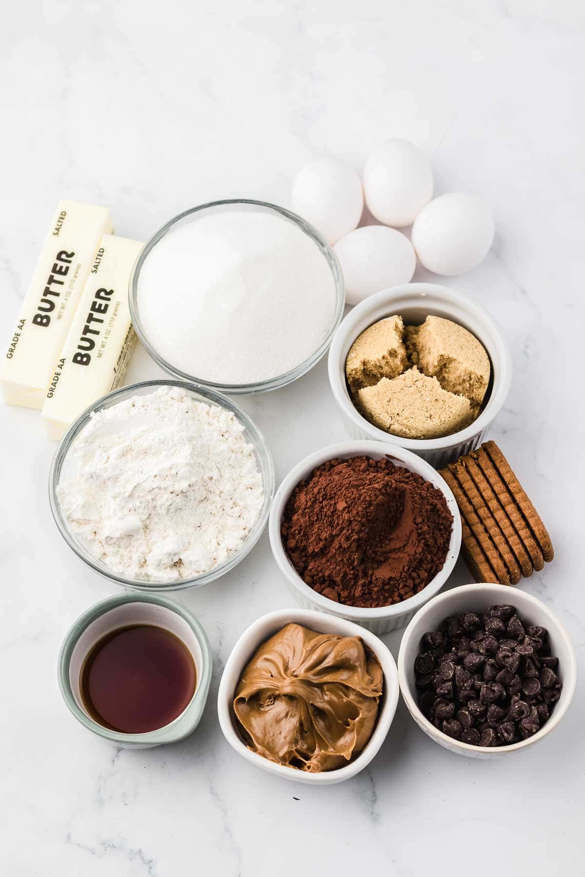 Baking ingredients on a white surface: eggs, butter, sugar, flour, cocoa, peanut butter, chocolate chips, vanilla, cookies.