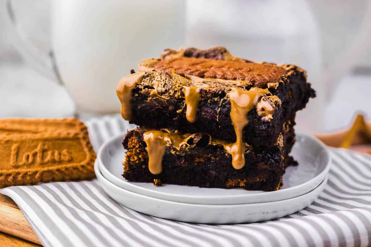 Two chocolate brownies with caramel and a Lotus biscuit on top, stacked on a plate next to a glass of milk.