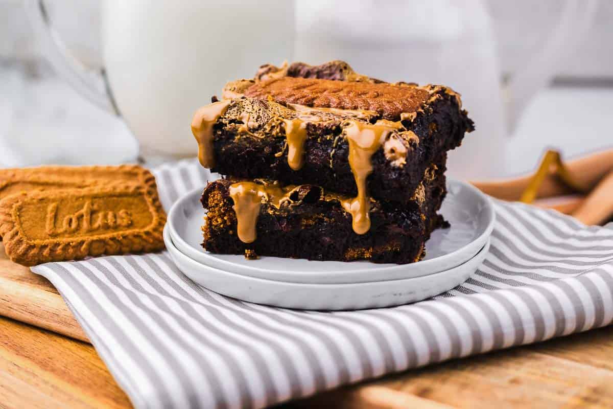 Two brownies topped with drizzled cookie butter and a biscuit, served on a plate with a glass of milk in the background.