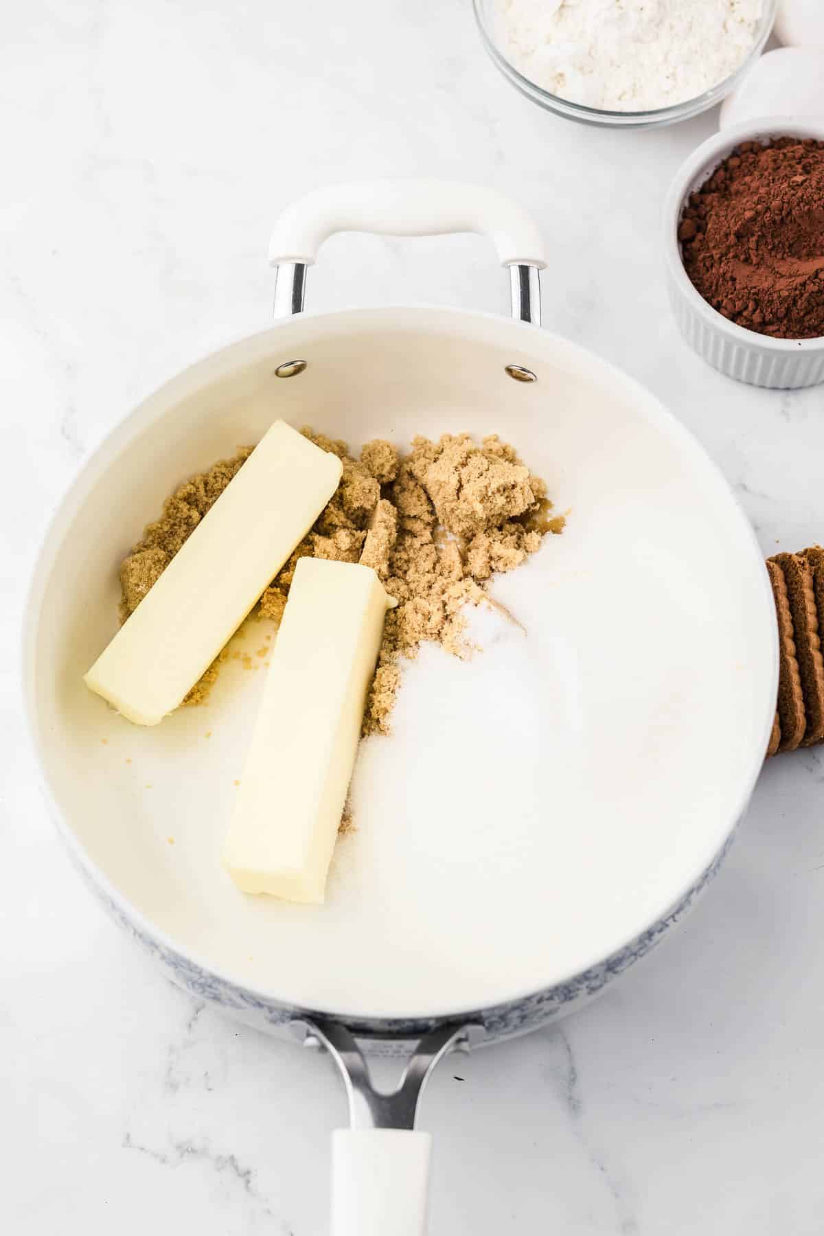 Two sticks of butter, brown sugar, and white sugar in a white pan; bowls of flour and cocoa nearby.