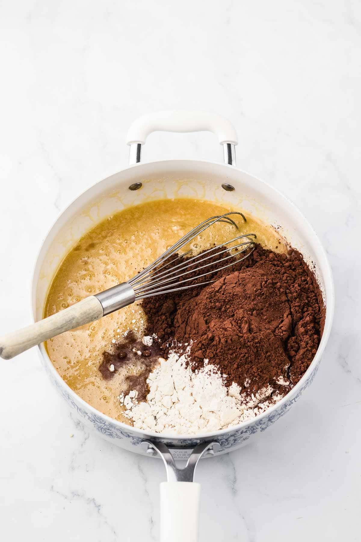A whisk in a bowl with melted butter, cocoa powder, and flour on a white marble surface.