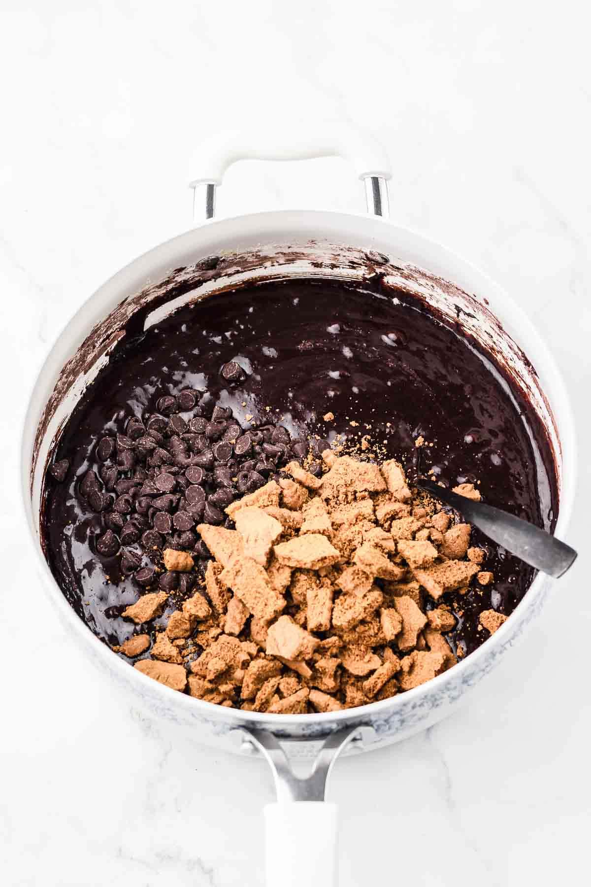 A pot of brownie batter with chocolate chips and crushed graham crackers being mixed with a spoon.