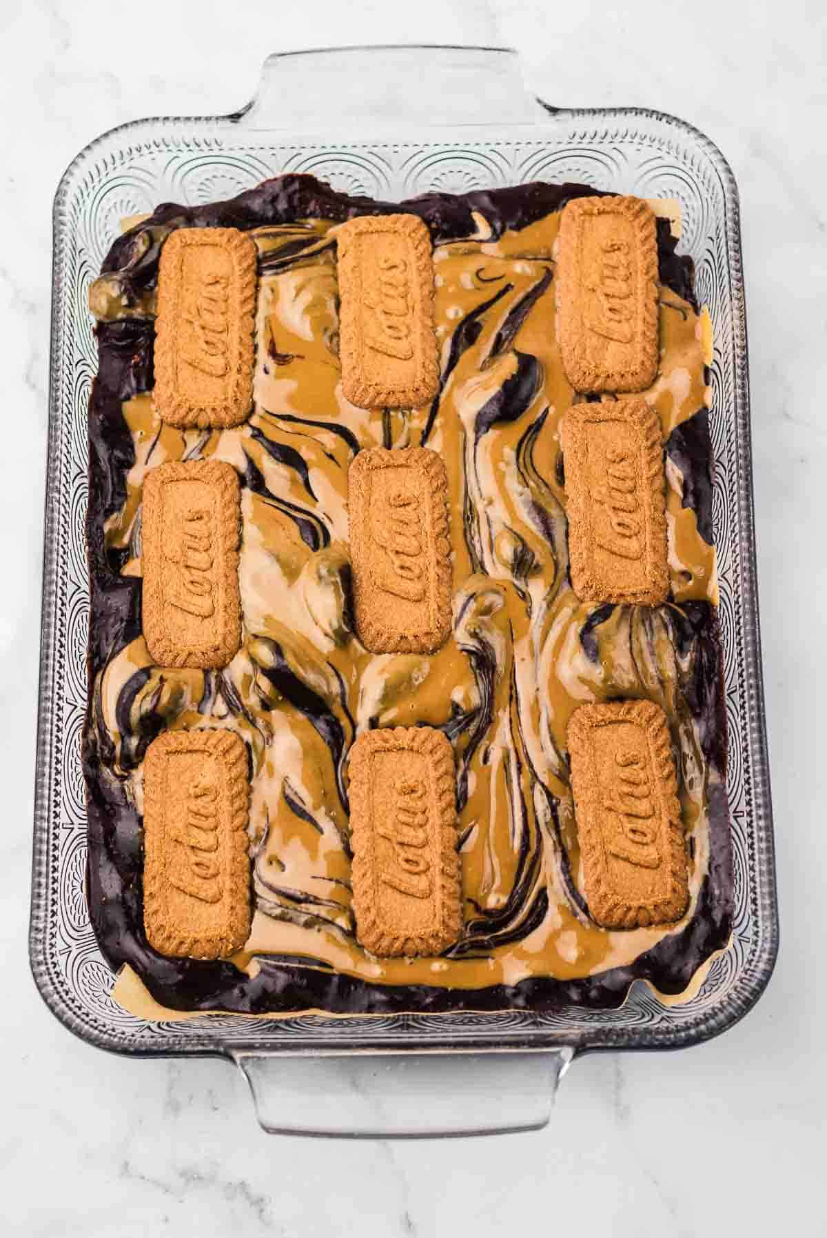 A glass baking dish with Biscoff Brownies, topped with cookie butter swirls and rectangular Biscoff cookies arranged on top.