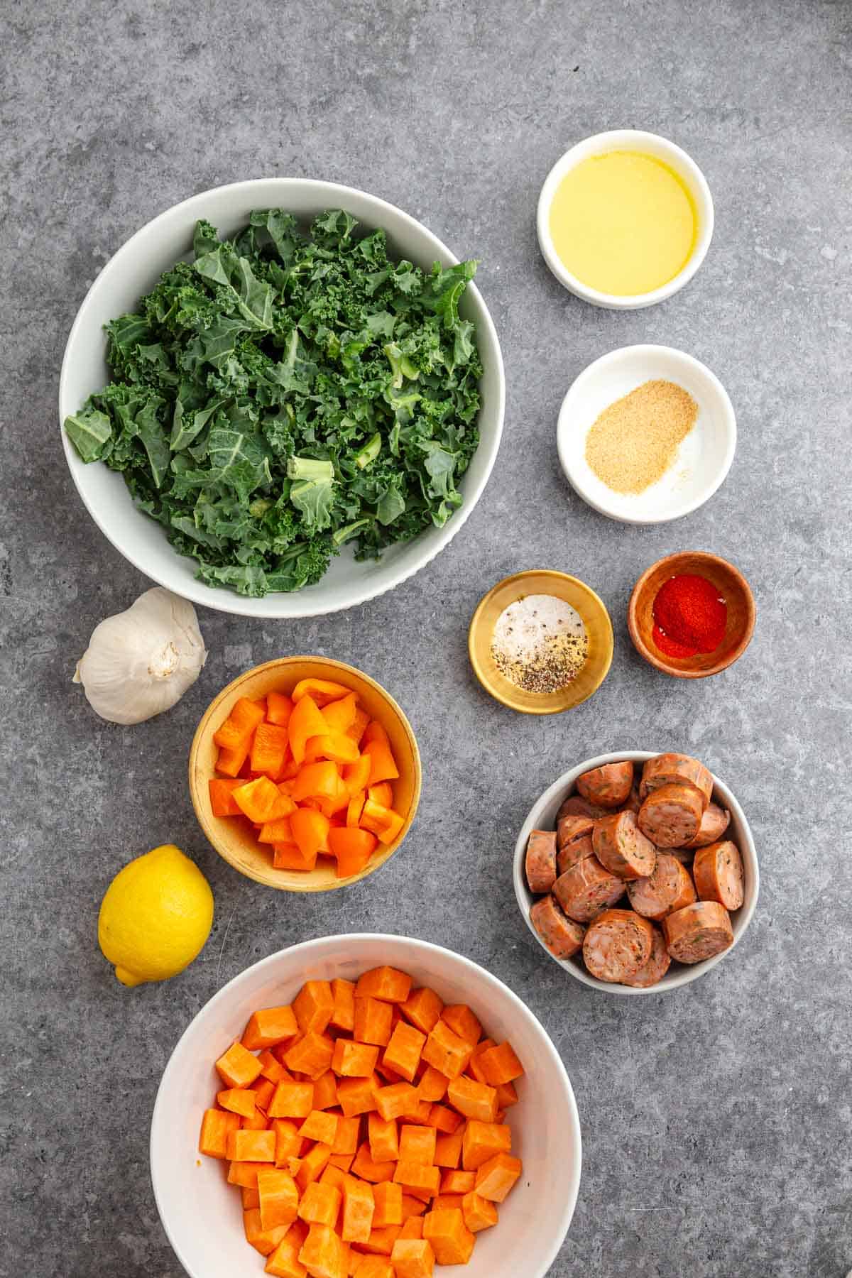 Bowls of chopped kale, sweet potatoes, sausage, bell pepper, and seasonings are arranged on a gray surface&mdash;perfect for an easy sheet pan meal.