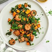 Sausage, sweet potatoes, and kale served on a white plate, garnished with fresh herbs and a sprinkle of pepper on the side.