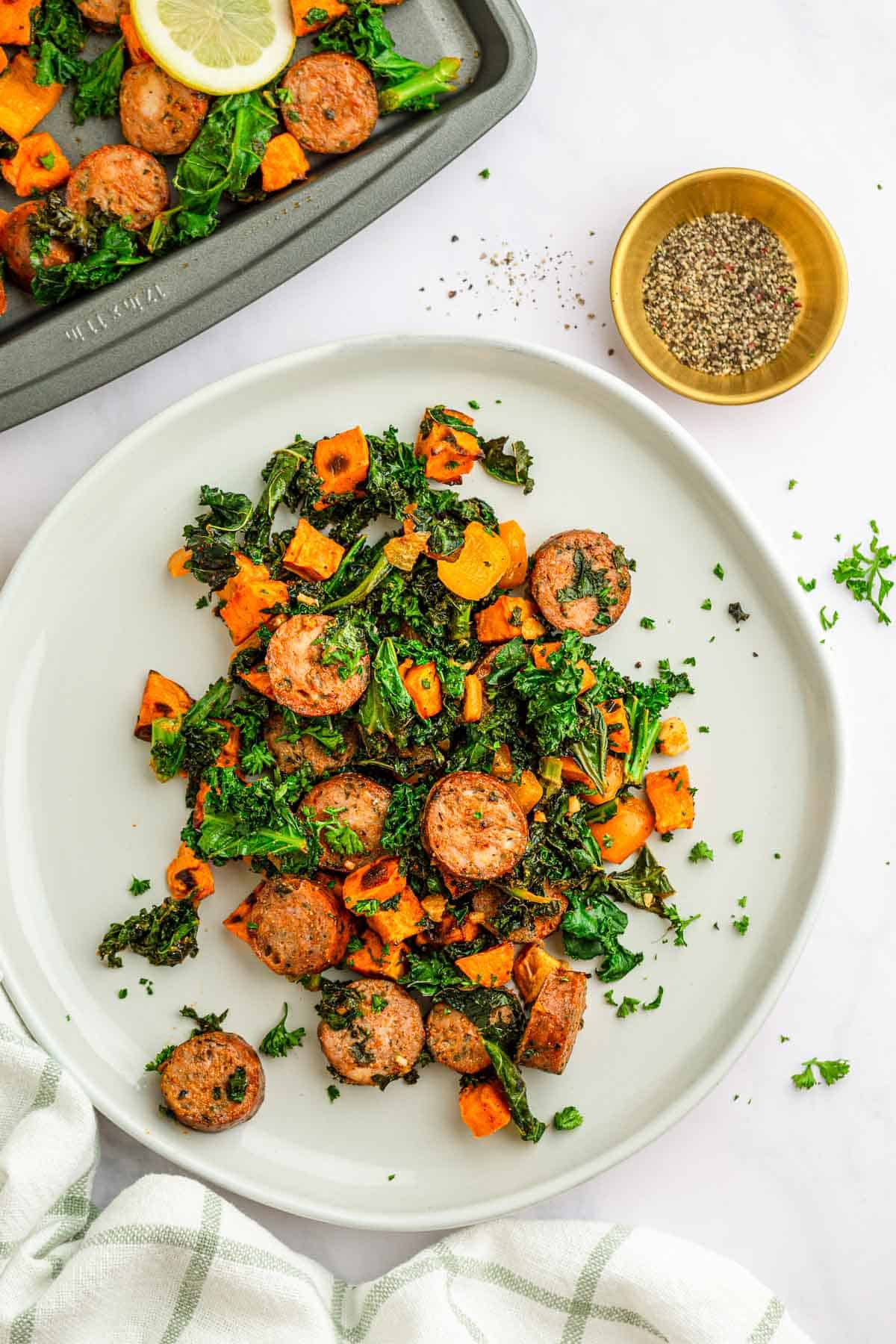 Sausage, sweet potatoes, and kale served on a white plate, garnished with fresh herbs and a sprinkle of pepper on the side.