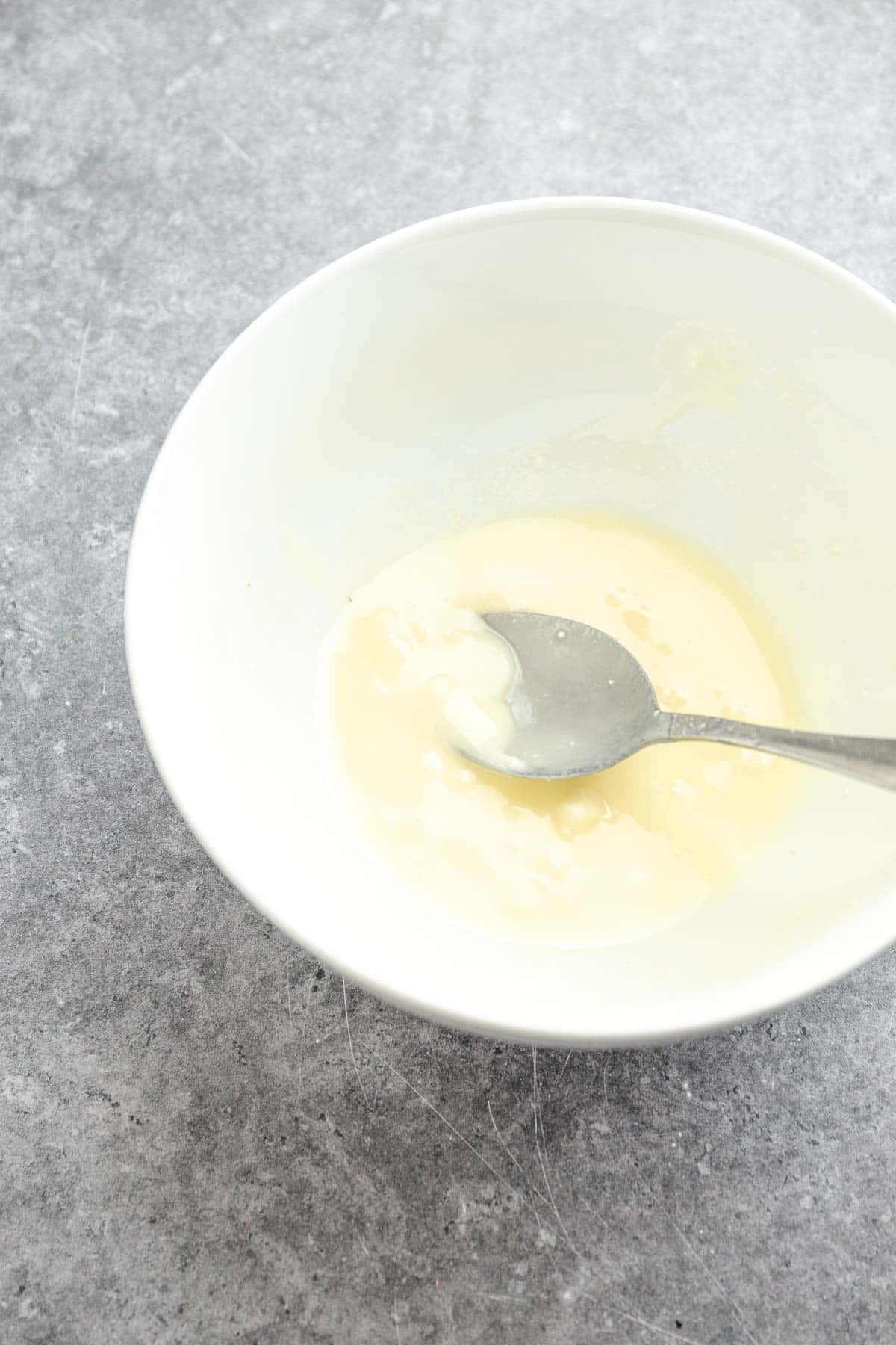 A metal spoon in a white bowl with a small amount of creamy white mixture on a gray surface, perfect for pairing with roasted sweet potatoes or sheet pan sausage dishes.