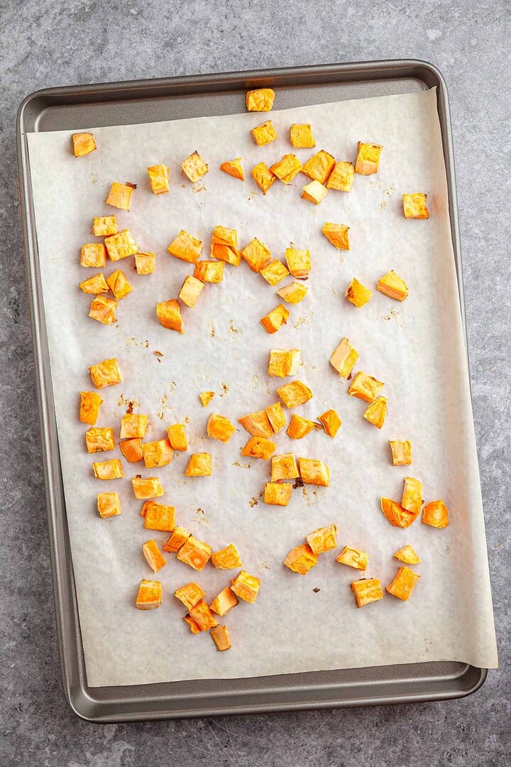 Diced roasted sweet potatoes and sausage on a parchment-lined sheet pan, spread out on a gray countertop.