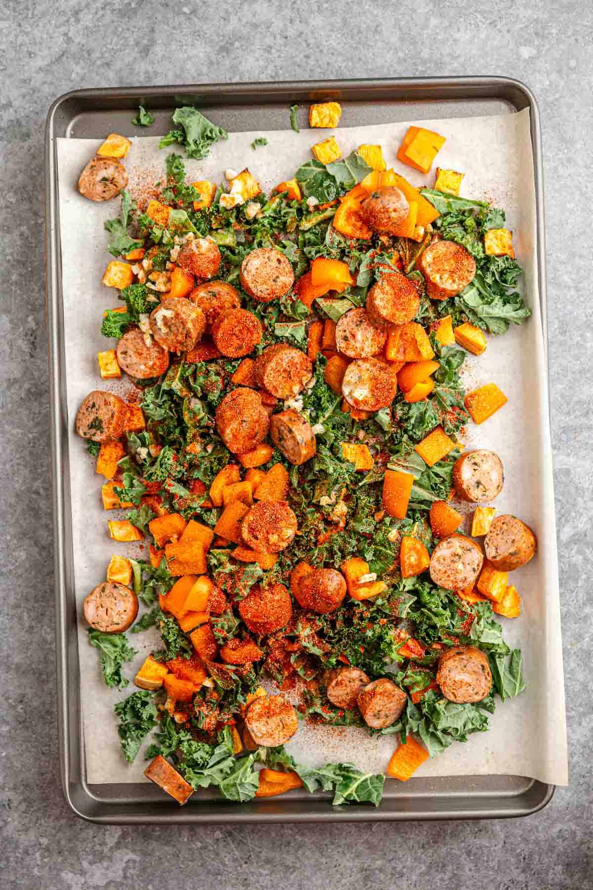 A sheet pan with sliced sausage, diced butternut squash, sweet potatoes, and kale, all seasoned and ready to bake.