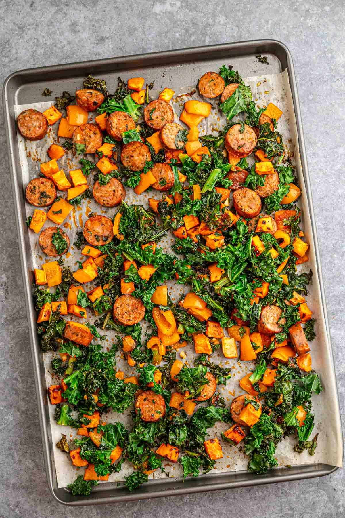 Sheet pan featuring roasted sweet potatoes, kale, bell peppers, and savory sausage slices on a gray surface.