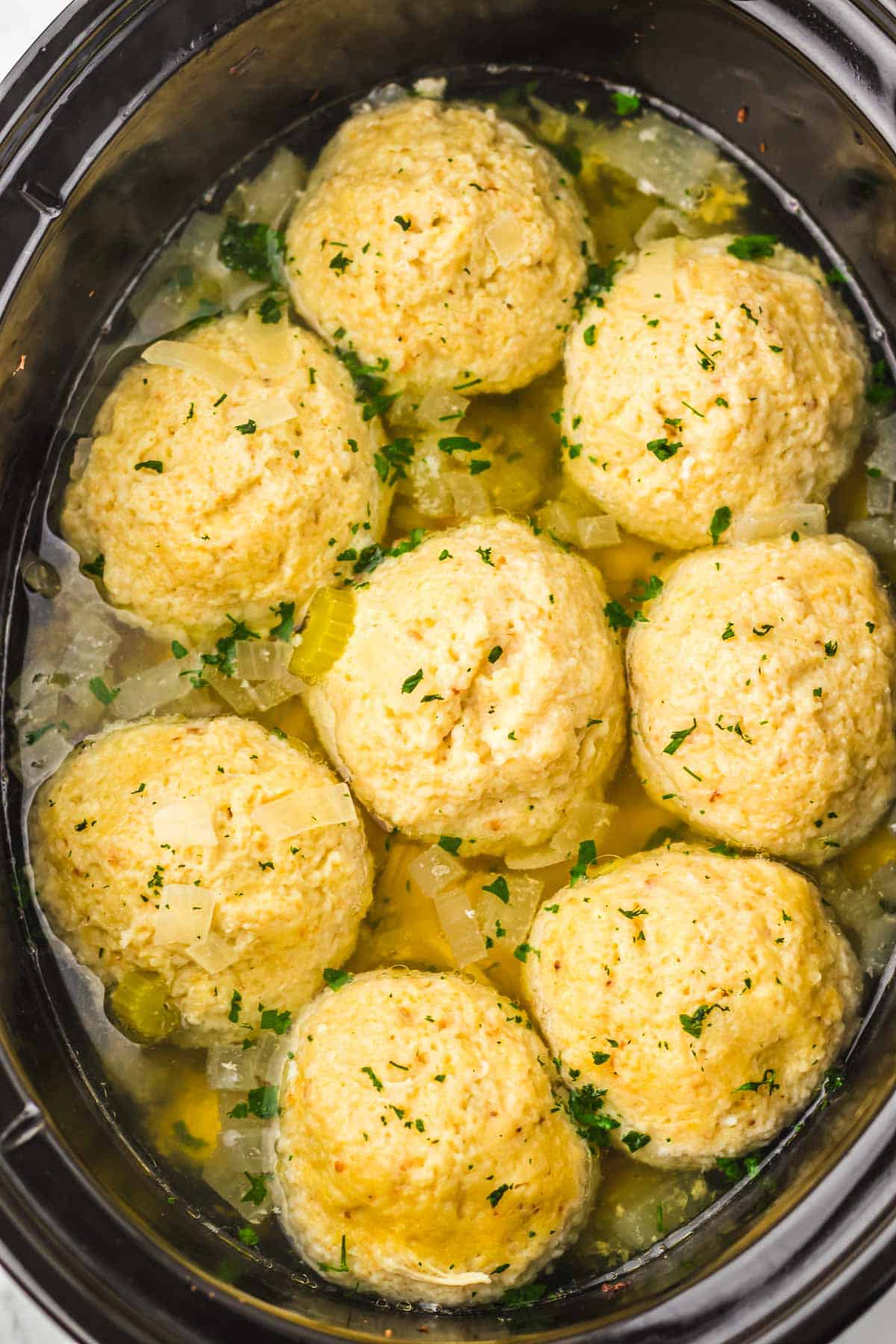 Seven matzo balls in broth with chopped onions and herbs simmer in a slow cooker, topped with fresh parsley.