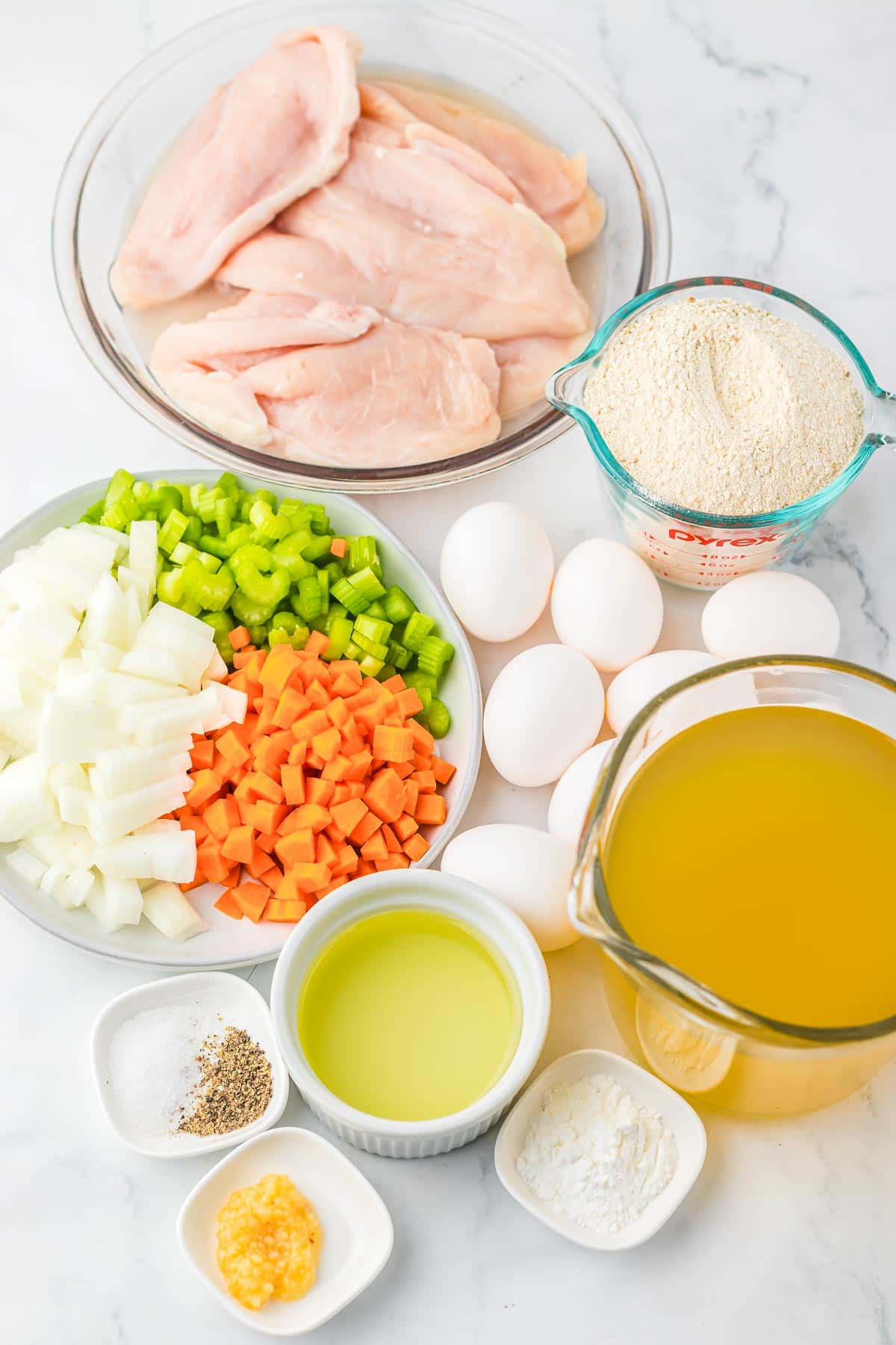 Raw chicken, chopped onions, carrots, celery, eggs, broth, oil, breadcrumbs, garlic, and seasonings on a white surface—perfect ingredients to start your favorite Matzo Ball Soup recipe.