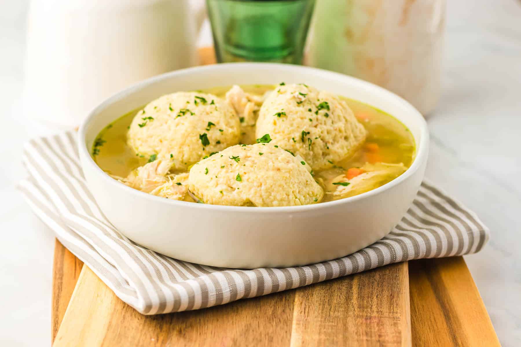 A bowl of Matzo Ball Soup featuring three matzo balls, chicken, and vegetables sits on a striped napkin and wooden board—perfect for trying a new recipe or making in your slow cooker.