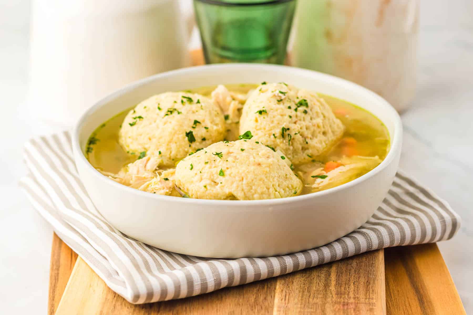A bowl of chicken and dumplings soup with three dumplings, on a striped cloth and wooden board—reminiscent of a classic matzo ball soup recipe.