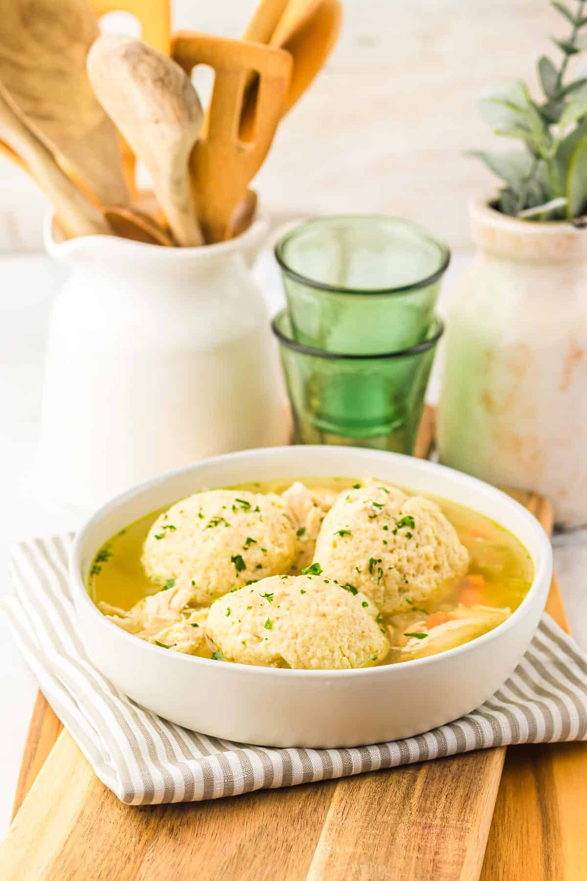 A bowl of chicken and dumplings soup, reminiscent of matzo ball soup, sits on a striped napkin, with wooden utensils and green glasses in the background—a comforting recipe perfect for the slow cooker.