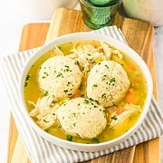 A bowl of chicken and dumplings soup, made from a comforting slow cooker recipe, sits on a striped napkin with glasses and jars in the background.