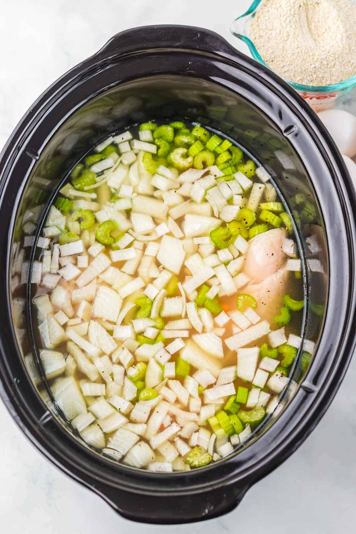 Slow cooker filled with raw chicken, chopped onions, and celery in broth—perfect for a comforting Recipe inspired by Matzo Ball Soup, with a measuring cup of dry quinoa nearby.