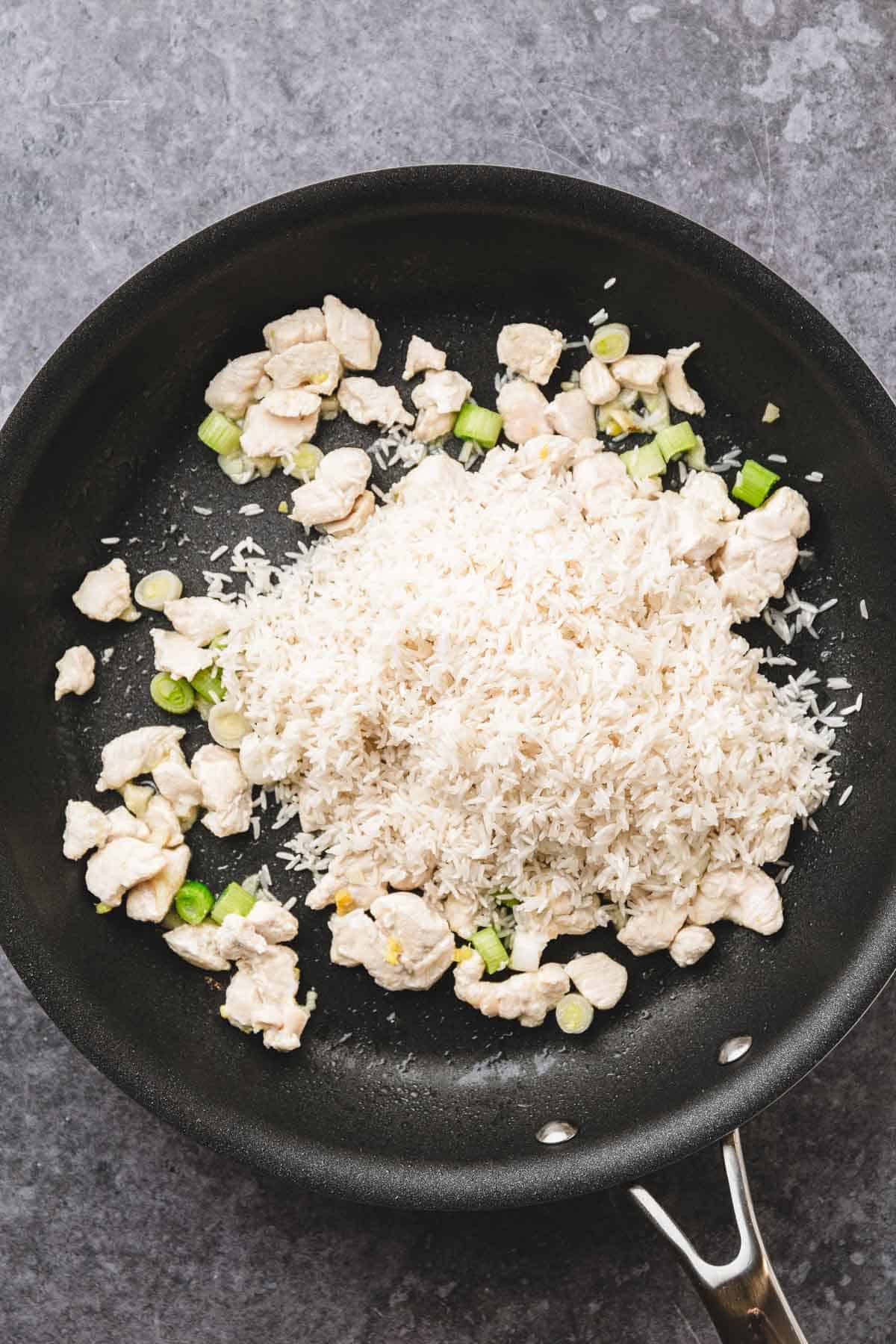 Raw rice, chopped chicken, and green onions in a black skillet on a gray countertop&mdash;perfect ingredients for a delicious teriyaki chicken fried rice recipe.