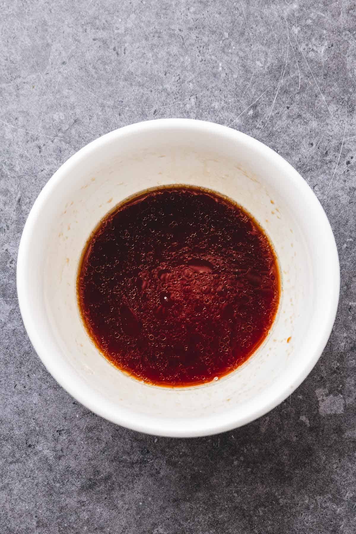 White bowl filled with a dark brown teriyaki chicken sauce, placed on a gray textured surface.