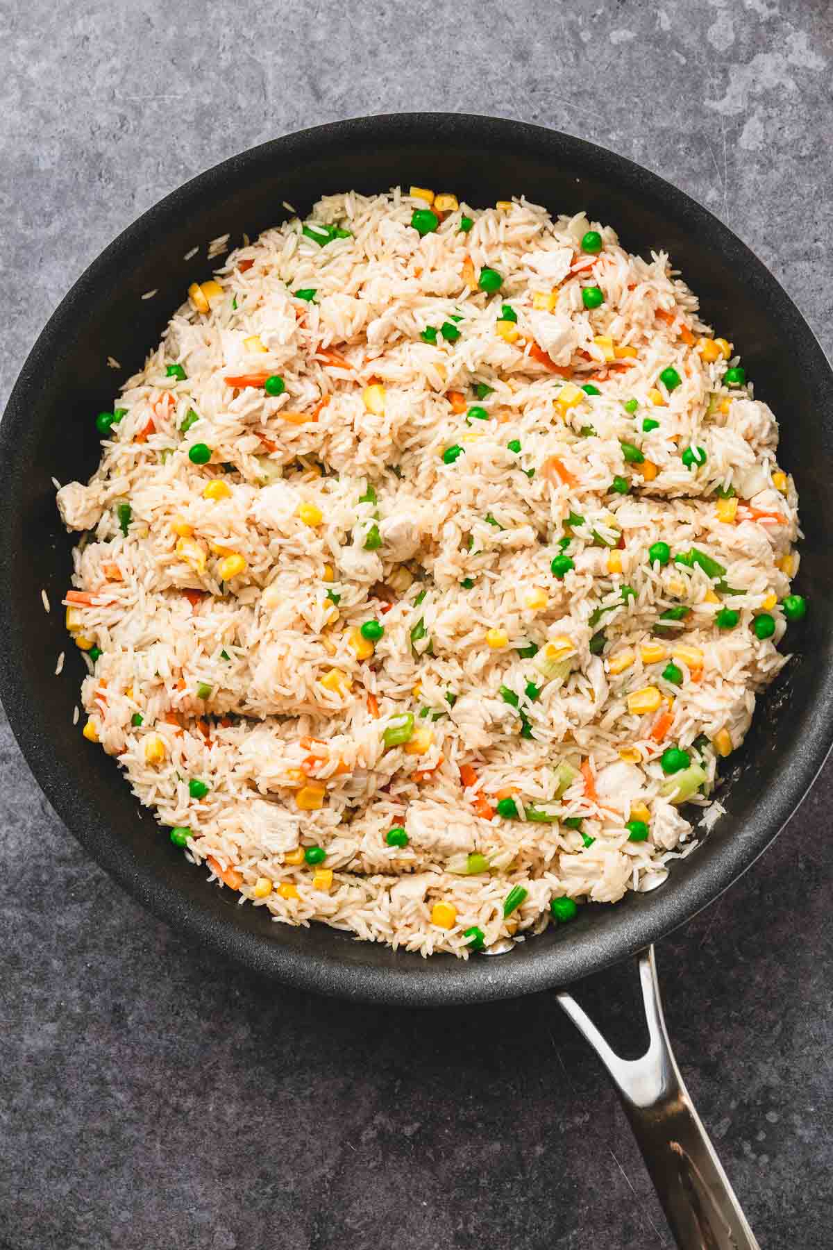 A skillet filled with fried rice, peas, corn, carrots, and shredded teriyaki chicken creates a flavorful recipe on a gray surface.