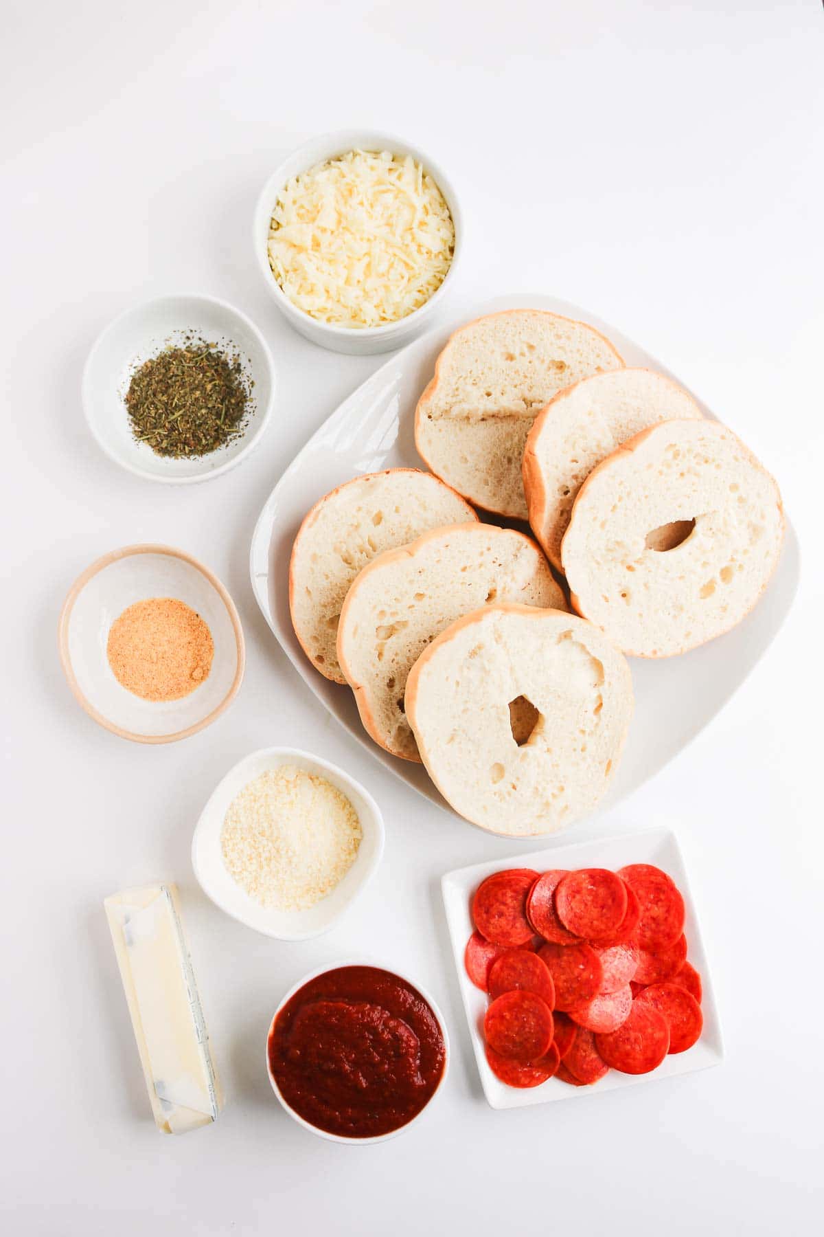 Sliced bagels, pepperoni, shredded cheese, pizza sauce, butter, herbs, and seasonings create delicious Homemade Pizza Bagels on a white background.